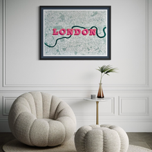 Load image into Gallery viewer, Framed, screen-printed London map wall art featuring a stylised pink 'LONDON' text design. Hung on a white panelled wall above a cream boucle armchair and small side table with a plant.