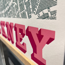 Load image into Gallery viewer, Close-up of Hackney map screen-print showing large pink block letters over a white background, with a dark map-style graphic above and a light wood edge visible.
