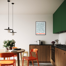 Load image into Gallery viewer, A framed, screen-printed street map print of De Beauvoir Town, London, hangs on a white wall in a modern kitchen and dining area. The print features a detailed map of the area with red and orange text at the bottom, surrounded by orange chairs, a wooden table, and dark wood cabinetry.