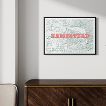 Load image into Gallery viewer, A framed art print featuring a vintage map of Hampstead with the word 'HAMPSTEAD' in large coral-pink letters, hanging on an off-white wall above a dark wood cabinet.