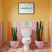 Load image into Gallery viewer, A framed screen-printed map print featuring the word 'HAMPSTEAD' in teal letters in a black frame, hanging on a mustard yellow wall above a white toilet in a brightly colored bathroom setting with pink, orange, and yellow tiles, flanked by two potted cacti in pink pots.
