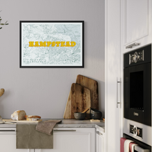 Load image into Gallery viewer, A framed art print featuring a vintage map of Hampstead with bold yellow text, displayed on a light gray kitchen wall above a countertop with wooden cutting boards and a loaf of bread.