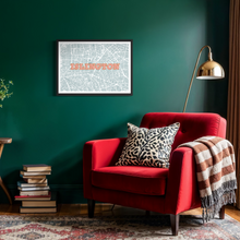 Load image into Gallery viewer, A framed map print of Islington with pink and gold lettering, displayed on a dark green wall above a red armchair with a patterned pillow and throw blanket in a styled room setting.