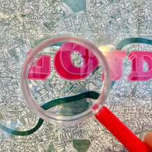 Load image into Gallery viewer, A clear magnifying glass with a red handle is held over a vintage-style map, focusing on bright pink letters that spell the word London.