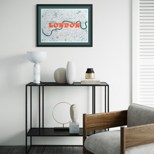 Load image into Gallery viewer, A framed art print featuring a London map with the word 'LONDON' in orange and the Thames in green, displayed above a black console table in a modern living room setting.

