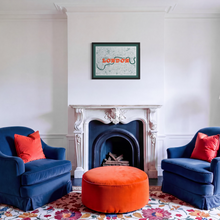 Load image into Gallery viewer, A framed London map screen print hangs above a white ornate fireplace in a styled living room. Two deep blue armchairs with bright orange pillows sit on either side of a round orange ottoman, all set on a colorful floral rug.
