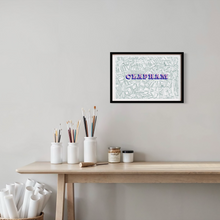 Load image into Gallery viewer, A framed screen-printed Clapham map with a blue and pink text with the word 'CLAPHAM', hanging on a light grey wall above a wooden table with white jars and art supplies.
