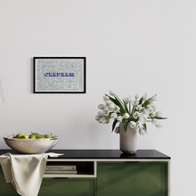 Load image into Gallery viewer, A framed vintage Clapham map, screen-printed in green with blue and pink text, with the word 'CLAPHAM', hanging on a white wall above a green cabinet with white tulips in a vase and a bowl of apples.ink text 
