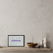Load image into Gallery viewer, A framed Clapham map screen print featuring the word 'CLAPHAM' in blue and pink text, displayed on a rustic wooden shelf against a textured grey wall alongside decorative objects.
