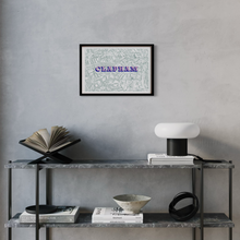 Load image into Gallery viewer, A framed screen-printed vintage Clapham map with pink and blue text, displayed on a grey wall above a modern grey console table with books and a lamp.
