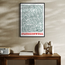 Load image into Gallery viewer, A framed street map poster of Clerkwenwell hangs on a textured beige wall above a wooden cabinet with decorative ceramic vases and books. The poster features a detailed light green map with the word 'CLERKENWELL' in bold orange letters at the bottom.
