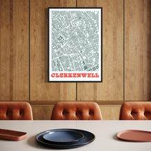 Load image into Gallery viewer, A framed street map print of Clerkenwell with a bold orange title band, displayed on a wood-panelled wall above a dining table with chairs and plates.
