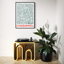 Load image into Gallery viewer, A framed screen-printed map poster of Clerkenwell printed in muted green-grey with bold orange text at the bottom, displayed on a wall above a yellow record stand with vinyl records next to a potted plant in a bright room.
