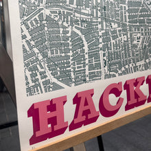 Load image into Gallery viewer, A close-up of a Hackney street map print with green street lines, featuring large pink block letters spelling 'HACKNEY' along the bottom, mounted on a wooden display stand.
