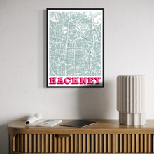Load image into Gallery viewer, A framed street map poster of Hackney hanging on a light gray wall above a wooden console table with a lamp, notebook, and tablet.
