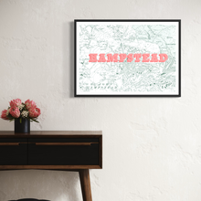 Load image into Gallery viewer, A framed vintage-style map print of Hampstead with bold pink 3D block letters spelling 'HAMPSTEAD' across the center, hanging on a white textured wall above a wooden console table with a vase of pink flowers.
