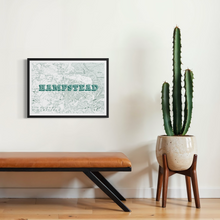 Load image into Gallery viewer, A framed screen-printed vintage-style map of Hampstead with the word 'HEMPSTEAD' in 3D block letters, displayed on a white wall in a styled room setting with a leather bench and a potted cactus.
